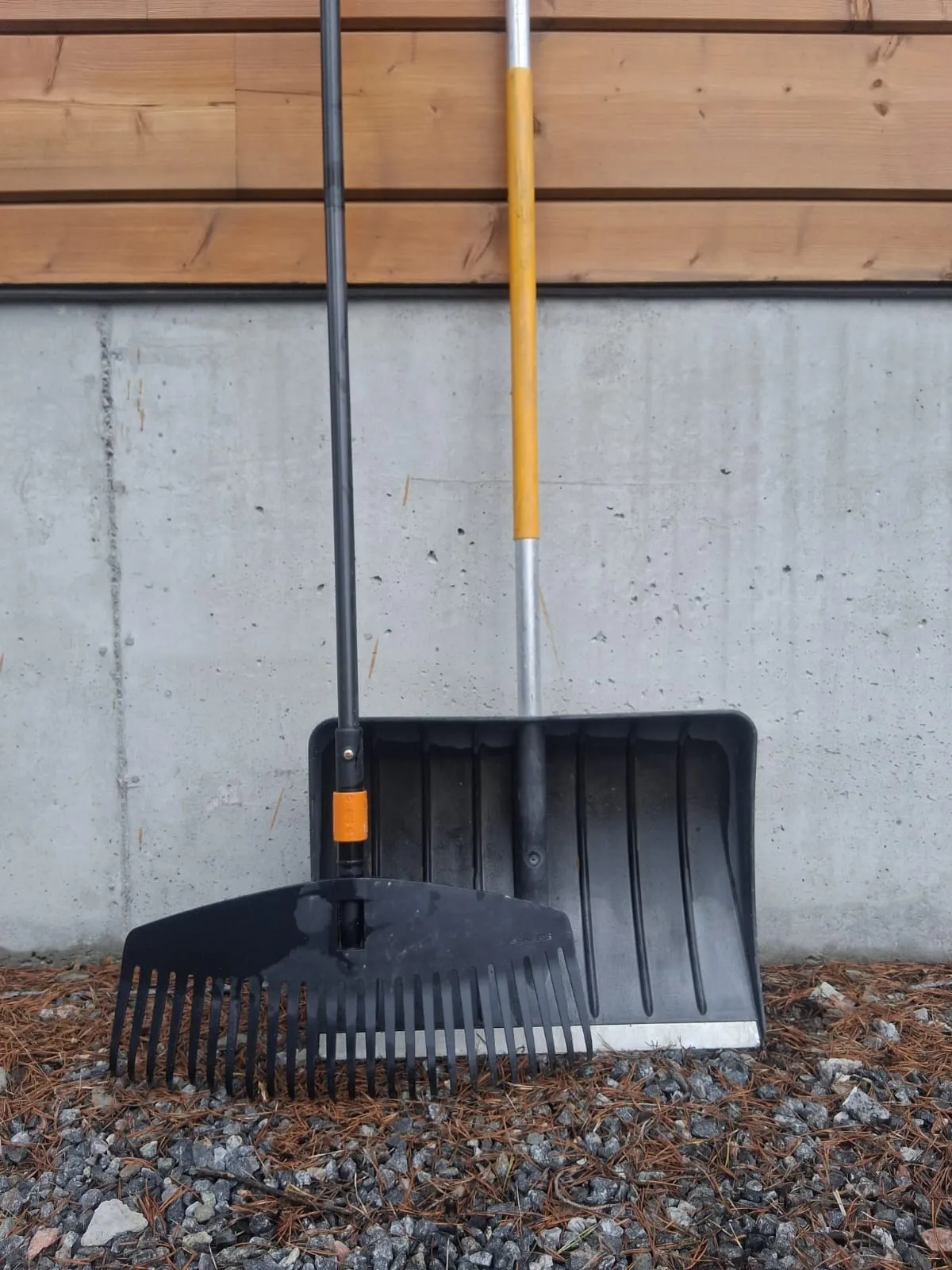 Kuva näyttää mustan haravan ja lumikolan nojaamassa harmaata seinää vasten, puinen seinä yläpuolella.