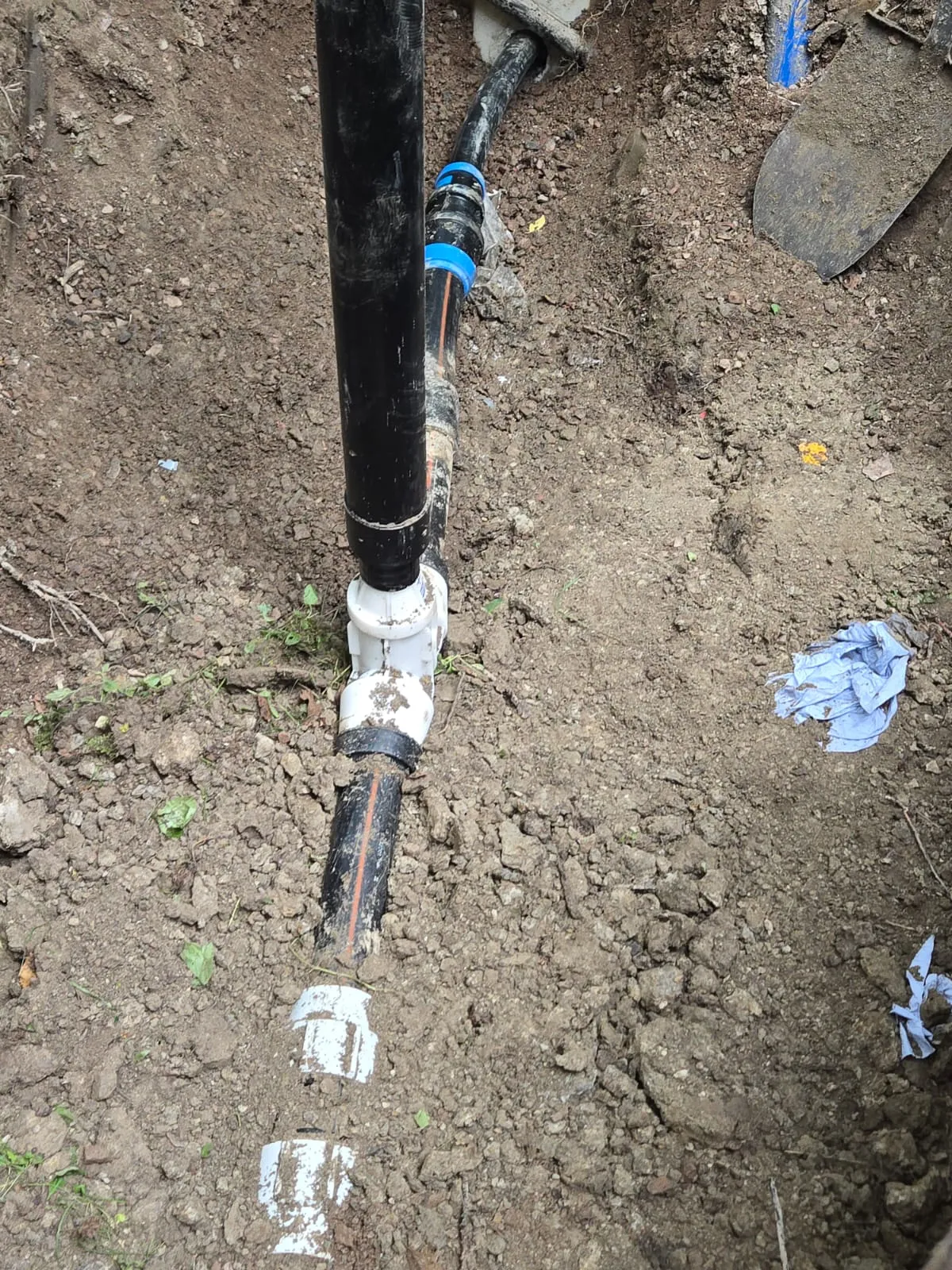 Maahan kaivettu kuoppa, jossa näkyy mustia ja valkoisia muoviputkia sekä lapio.