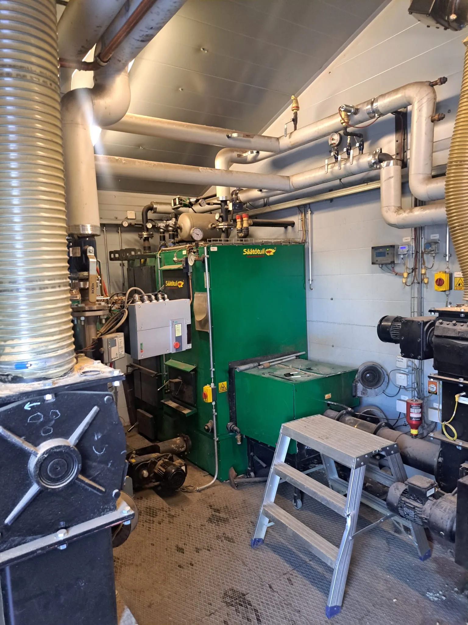 Sisätilassa on vihreä lämmityskattila, metalliputkia katossa, harmaa ohjauspaneeli ja alumiininen porrasjakkara. Valoisa tila.