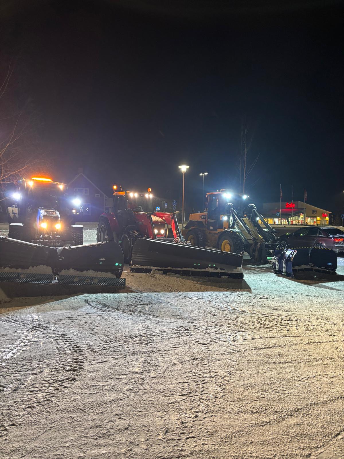 Yöllinen kuva lumiauroista parkkipaikalla, taustalla valaistu kauppa ja katuvalot.