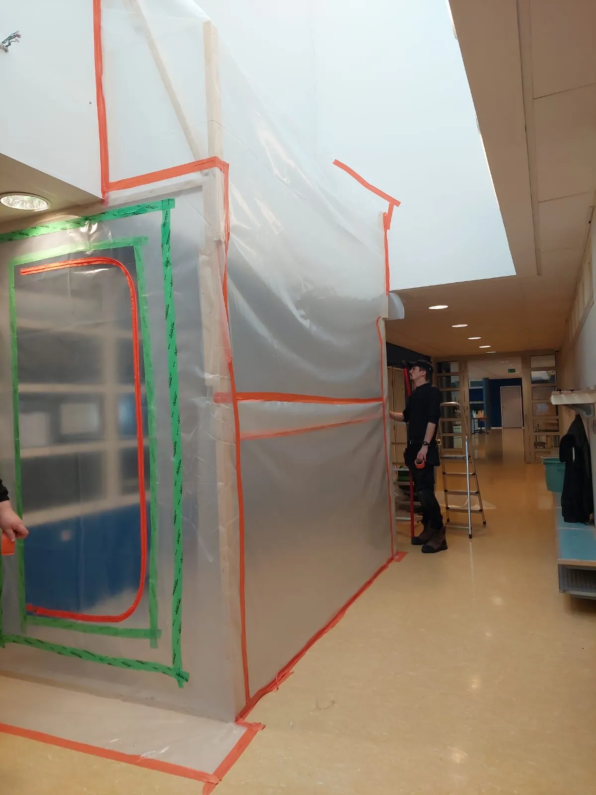 Kuvassa näkyy muovilla ja teipillä suojattu alue käytävällä, jossa mies seisoo tikkaiden vieressä. Valoisa käytävä, jossa on valkoiset seinät ja kattoikkuna.