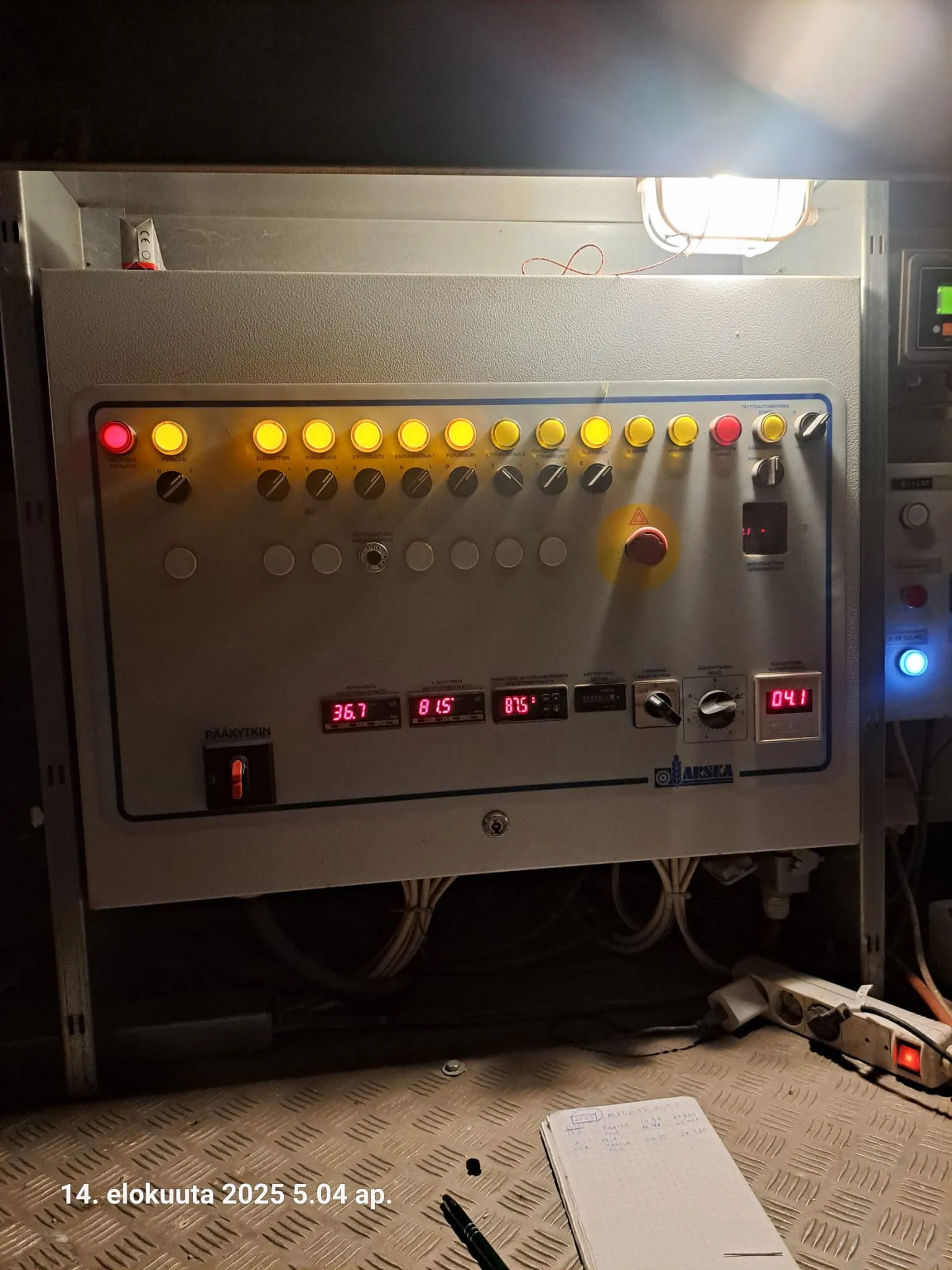 Ohjauspaneeli, jossa on valoja, painikkeita ja näyttöjä, sekä muistiinpanovälineet metallipinnalla.