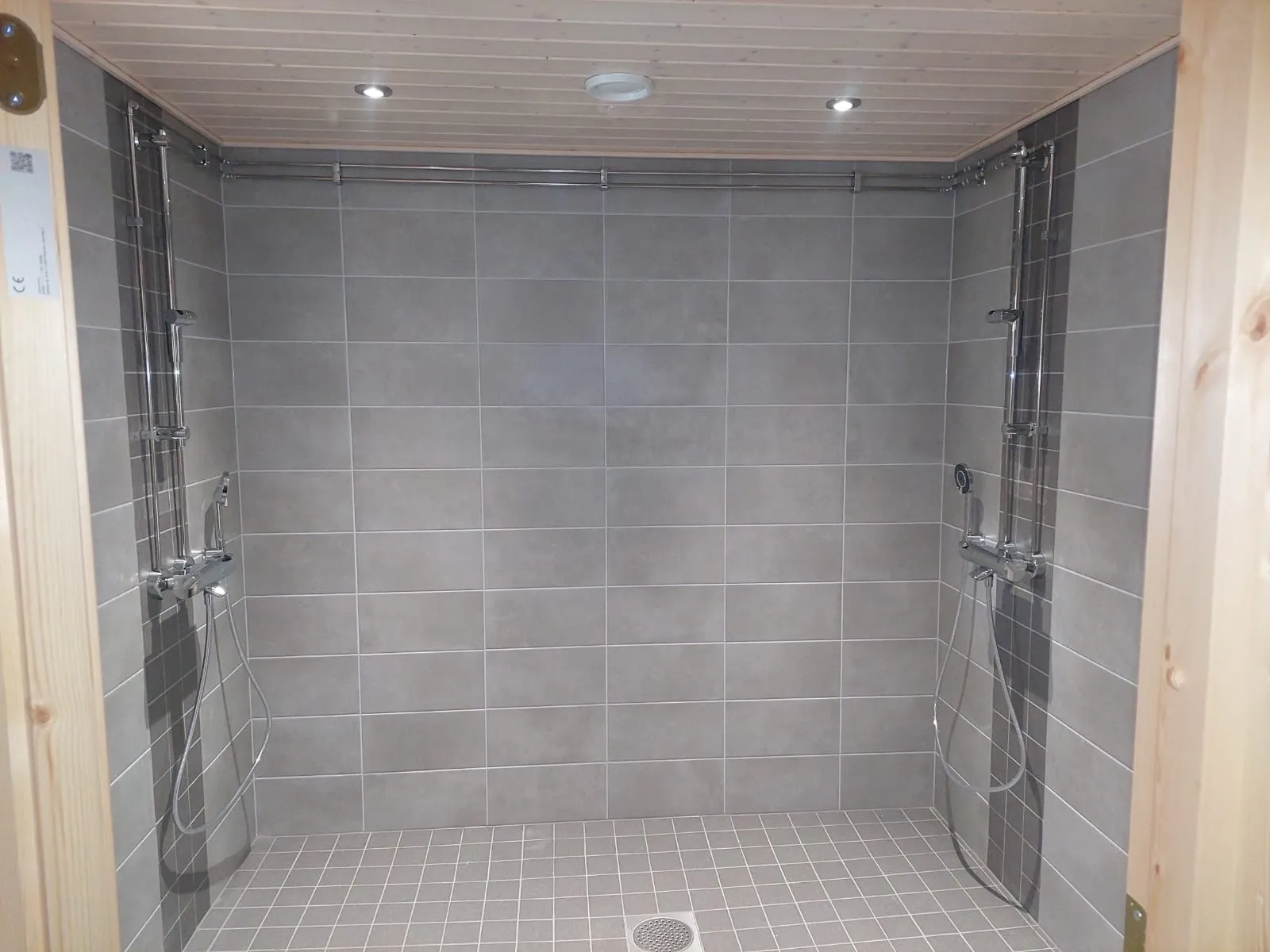 Harmaa suihkuhuone, jossa on kaksi suihkua, laattalattia ja puinen katto, valaistus on kirkas.