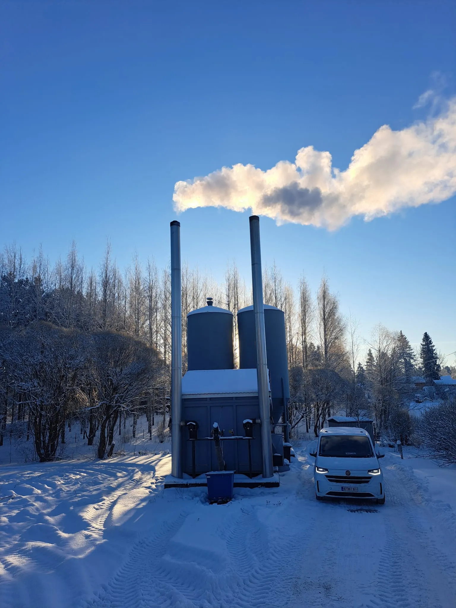 Talvinen maisema, jossa näkyy lumipeitteinen maasto, tehdasrakennus savupiippuineen ja valkoinen pakettiauto. Taustalla sininen taivas ja pilviä.