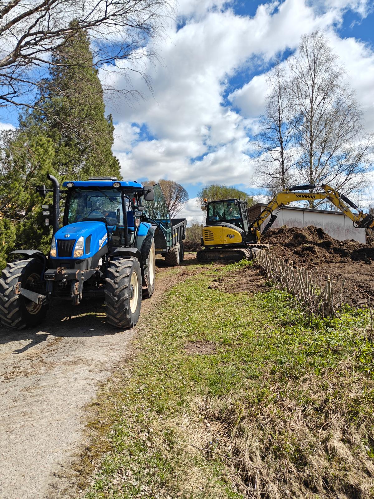 Aurinkoisessa maisemassa on sininen traktori peräkärryineen ja keltainen kaivinkone, jotka ovat maataloustöissä. Taustalla näkyy puita ja sininen taivas pilvineen.