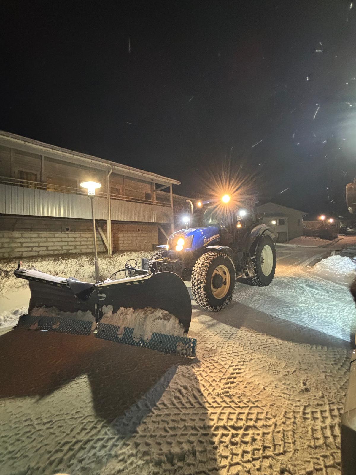 Kuvassa on New Holland -merkkinen traktori, jossa on lumiaura kiinnitettynä, lumisella tiellä yöllä. Traktorin valot ovat päällä, ja taustalla näkyy rakennuksia ja katuvaloja.